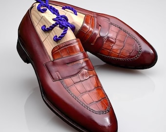 Könnte beinhalten: Ein Paar braune Leder-Loafer mit krokodiltexturierter Vorderkappe und Zehen. Die Schuhe haben ein poliertes Finish und werden mit hölzernen Schuhspannern und lila Seil präsentiert. Die Loafer sind ein klassischer Stil.
