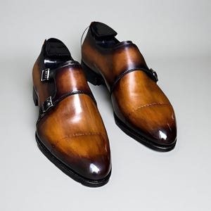Pode incluir: Um par de sapatos monk strap de couro castanho com acabamento brilhante. Os sapatos apresentam um fecho de fivela dupla e um detalhe castanho escuro à volta do tornozelo. As solas são pretas.