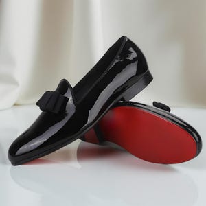 May include: Pair of black patent leather loafers with a bow detail. The shoes have a glossy finish and a red sole. The loafers are displayed on a reflective surface.