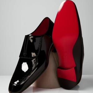 May include: Black patent leather dress shoes with a glossy finish. One shoe is upright, showcasing the design, while the other reveals a vibrant red sole. The shoes have a classic, formal style, suitable for special occasions.