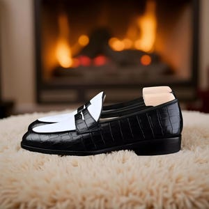 Op de afbeelding: Een paar zwart-witte leren loafers met een krokodillenstructuur. De schoenen hebben een klassiek ontwerp met een witte wreef en een zwarte body. De schoenen worden gepresenteerd op een zacht, cr&egrave;mekleurig tapijt.