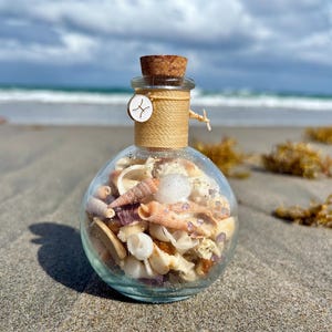 May include: A clear glass bottle filled with seashells, small purple crystals, and sand, sealed with a cork and twine. A small white charm with a symbol is attached to the twine. The bottle sits on a sandy beach with the ocean in the background.
