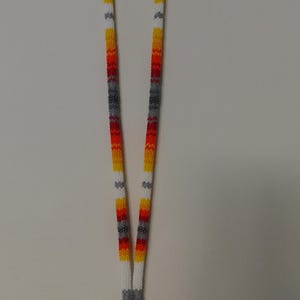 Peut inclure: Un long cordon porte-badge composé de petites perles dans un motif répétitif blanc, jaune, orange, rouge et gris. Le cordon perlé forme une boucle à la base, terminée par un anneau métallique argenté et un fermoir mousqueton.
