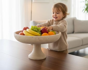 May include: A beige pedestal fruit bowl filled with fresh fruit, including red apples, green grapes, bananas, and oranges. A young child is reaching for the fruit. The bowl sits on a dark brown wooden table.