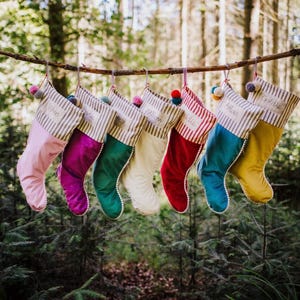 May include: A selection of festive Christmas stockings suspended from a branch. The stockings are in various colours: pink, purple, green, cream, red, teal, and yellow. Each stocking features a striped top and is personalised with a name.
