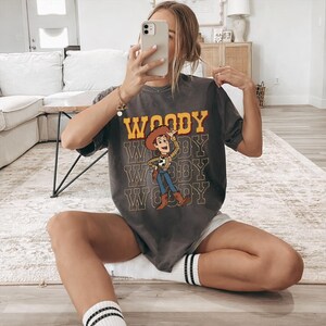 Pode incluir: T-shirt cinzenta escura com a palavra "WOODY" em letras amarelas e uma imagem de desenho animado de Woody de Toy Story. A camisa tem um visual desgastado e &eacute; combinada com cal&ccedil;&otilde;es brancos e meias listradas.