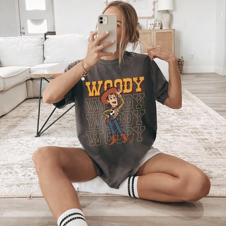 Pode incluir: T-shirt cinzenta escura com a palavra "WOODY" em letras amarelas e uma imagem de desenho animado de Woody de Toy Story. A camisa tem um visual desgastado e &eacute; combinada com cal&ccedil;&otilde;es brancos e meias listradas.