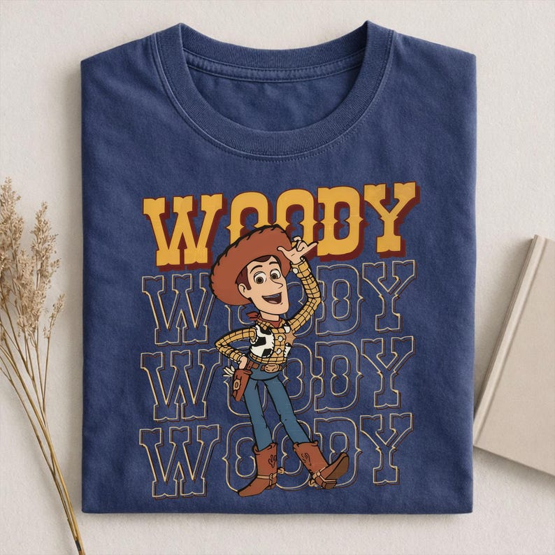 Pode incluir: Camiseta azul marinho com o personagem Woody de Toy Story. O design inclui a palavra "WOODY" em letras amarelas e vermelhas acima de uma ilustra&ccedil;&atilde;o de Woody. A palavra "WOODY" &eacute; repetida em um padr&atilde;o empilhado abaixo.