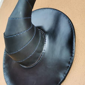 Könnte beinhalten: Ein schwarzer Hexenhut mit breiter Krempe und spitzer Krone. Der Hut besteht aus glänzendem Material und hat hellblaue Nähte an den Nähten und Kanten. Ein klassisches Halloween-Kostümzubehör.