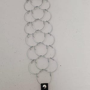 Pode incluir: Uma pulseira de cota de malha de metal prateado com uma correia e fivela de couro preto. A cota de malha é feita de anéis interligados. A pulseira é mostrada em uma superfície branca.