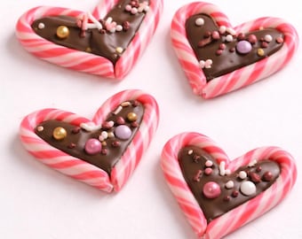 May include: Four heart-shaped candies with a chocolate filling and colorful sprinkles. Each candy is outlined with a red and white striped candy cane. The candies are on a white surface.