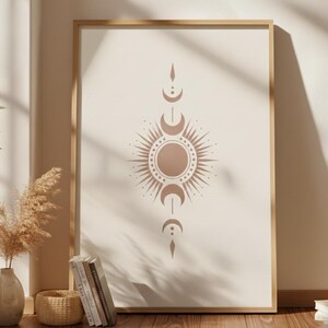 May include: A framed print featuring a celestial design with a sunburst and crescent moon motifs in a warm, earthy tone. The artwork is displayed in a light-colored wooden frame, leaning against a wall. The overall aesthetic is bohemian and minimalist.