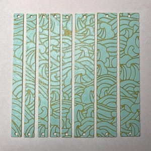 May include: Nine rectangular teal bookmarks with a gold wave pattern. Each bookmark has a small hole at the top. The wave design is a repeating pattern. The bookmarks vary slightly in length. The measurements are in centimetres.