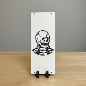Peut inclure: Panneau rectangulaire blanc avec une illustration noire d'un crâne et d'un col décoratif. Le panneau est soutenu par un support noir. L'illustration du crâne est détaillée avec des ombres et des lignes, lui donnant une esthétique vintage ou gothique.