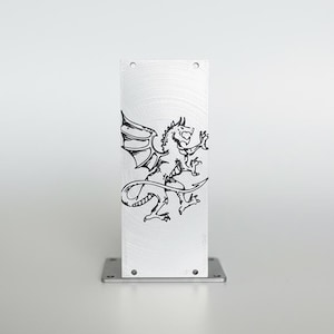 May include: A rectangular, brushed metal object with a black dragon illustration. The dragon is depicted with wings, a long tail, and outstretched claws. The object is mounted on a square metal base with four screw holes.