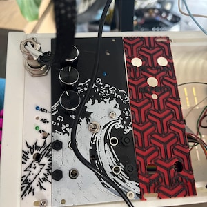 May include: A modular synthesizer panel featuring a black and white graphic of a crashing wave, three black control knobs, a metal toggle switch, and several input jacks. To the right is a red panel with a repeating geometric pattern and text.