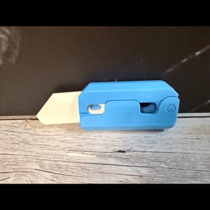 Könnte beinhalten: Ein blaues Universalmesser mit einer weißen Klinge. Das Messer besteht aus Kunststoff und hat einen Schiebemechanismus. Die Klinge ist teilweise ausgefahren und zeigt ihre scharfe Kante. Es liegt auf einer Holzoberfläche.