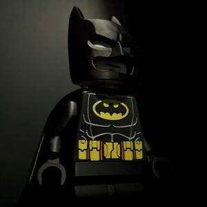 May include: A black Lego Batman figure with a yellow bat symbol on the chest. The figure has a black cape and a detailed design, including a yellow utility belt. The figure is a collectible item.