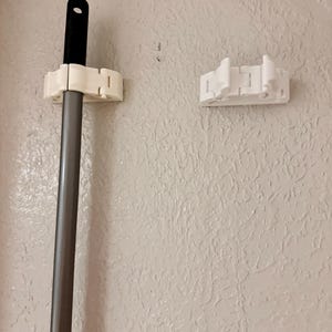May include: A white plastic wall-mounted mop and broom holder. The holder is designed to grip the handle of a mop or broom. The image shows the holder with a mop in place and a separate holder. The wall is painted a light beige colour.