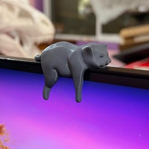 以下が含まれることがあります： コンピューターモニターの端に掛けるように設計された、グレーの3Dプリントされた猫の置物。猫はリラックスした姿勢で、脚が垂れ下がっています。背景には、紫と青のグラデーションのコンピューター画面が表示されています。