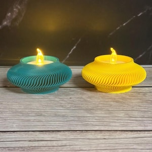 May include: Two decorative teal and yellow LED candle holders with a wavy design. Each candle holder has a flickering flame. The candle holders are on a wooden surface.