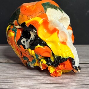 May include: A colorful, abstract skull sculpture. The skull is painted with vibrant hues of orange, yellow, black, and white, creating a unique and eye-catching design. The artwork is displayed on a wooden surface.