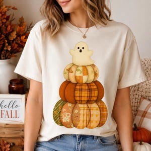 Peut inclure: Un t-shirt à manches courtes de couleur crème arborant un motif représentant une pile de citrouilles aux motifs et couleurs automnales variés, incluant l'orange, le jaune et le vert. Un petit fantôme clair à l'air surpris est posé au sommet.