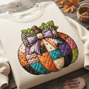 May include: A cream-colored crewneck sweatshirt featuring a large, quilted-style pumpkin graphic. The pumpkin is composed of various patterned fabric patches in orange, teal, purple, and black, topped with a decorative checkered bow.