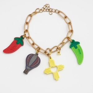 May include: Gold-tone charm bracelet featuring a red chili pepper, a green chili pepper, a hot air balloon, and a yellow cross-shaped charm. The bracelet has a chain link design.