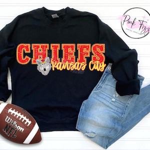 Peut inclure: Sweat-shirt noir avec le mot "CHIEFS" en lettres rouges et jaunes, et un loup de dessin animé. Les mots "Kansas City" sont en écriture jaune. Un ballon de football marron et blanc et un jean bleu sont également dans l'image.