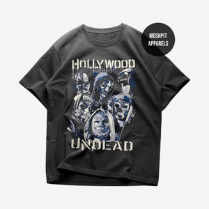 Può includere: T-shirt grigio scuro con un design grafico con le parole "HOLLYWOOD UNDEAD" e immagini di figure mascherate in tonalità blu e bianco. La maglietta ha un girocollo e maniche corte. Il logo del marchio "MOSHPIT APPARELS" è in alto a destra.