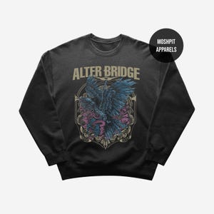 Op de afbeelding: Donkergrijze crewneck sweatshirt met de bandnaam "ALTER BRIDGE" in bruine letters boven een afbeelding van een raaf met blauwe vleugels en een roze bloemenontwerp. Het merk "MOSHPIT APPARELS" staat in een zwarte cirkel rechtsboven.