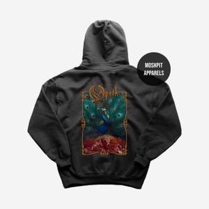 Puede incluir: Sudadera con capucha gris oscuro con un diseño de pavo real con plumas turquesas y azules, sobre un fondo rojo y naranja. La palabra "Opeth" está en oro sobre el pavo real. La sudadera también tiene el texto "MOSHPIT APPARELS".