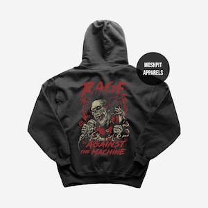 May include: Dark grey hoodie with a red and beige skull graphic holding a microphone. The word "RAGE" is above the skull, and "AGAINST THE MACHINE" is below. The hoodie has a hood and a "MOSHPIT APPARELS" logo.
