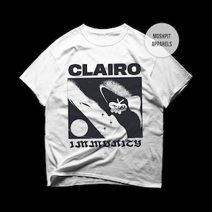 May include: White t-shirt with the word "CLAIRO" above a black and white graphic. The graphic includes a moon, stars, and a stylized face, with the word "IMMUNITY" below.