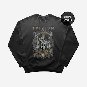 May include: Dark gray sweatshirt featuring the band name "TRIVIUM" in gold lettering above a detailed graphic. The graphic depicts three skeletons, flames, and ornate details. A black circle with "MOSHPIT APPARELS" is in the upper right corner.