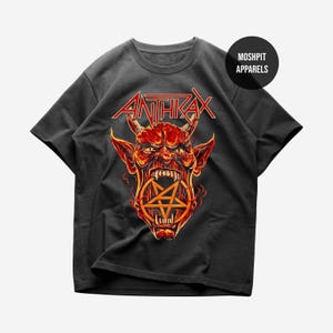 May include: Dark grey t-shirt with the band name "ANTHRAX" in red above a detailed illustration of a red demon with horns and a pentagram. The shirt has a crew neck and short sleeves.