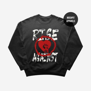 May include: Black crewneck sweatshirt with the words "RISE AGAINST" in white, with a red heart and fist graphic. The sweatshirt is a dark color, and the text and graphic are prominently displayed. The brand name "MOSHPIT APPARELS" is in a black circle.