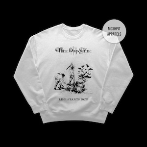 May include: White sweatshirt featuring a black graphic design with the text "Three Days Grace" and "Life Starts Now." The design depicts figures and bats. The brand name "Moshpit Apparels" is also visible.