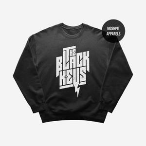 Könnte beinhalten: Ein schwarzes Sweatshirt mit dem weißen Schriftzug "THE BLACK KEYS" in einer kräftigen, stilisierten Schriftart und einem Blitz-Grafik. Das Sweatshirt hat auch einen kleinen, schwarzen Kreis mit dem Text "MOSHPIT APPARELS".