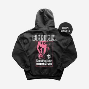 Może przedstawiać: Czarna bluza z kapturem z napisem "BIKINI KILL" nad różową grafiką. Grafika zawiera postać i słowa "REVOLUTION GIRL STYLE NOW". Okrągłe logo "MOSHPIT APPARELS" znajduje się w prawym górnym rogu.