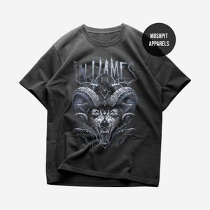 May include: Dark gray t-shirt featuring a detailed graphic of a snarling wolf with large horns and the text "IN FLAMES" in a stylized font. The shirt also has the text "MOSHPIT APPARELS" on a black circle.
