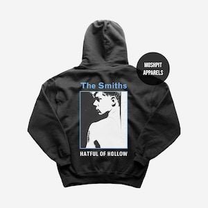 Op de afbeelding: Zwarte hoodie met een afbeelding van een mannenprofiel in een blauw omrand rechthoek, met de tekst "The Smiths" erboven en "Hatful of Hollow" eronder. Een cirkelvormig logo met "Moshpit Apparels" staat rechtsboven.