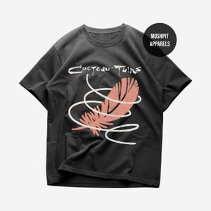 Peut inclure: T-shirt noir avec l'inscription "Cocteau Twins" en blanc au-dessus d'un motif de plume corail avec des spirales blanches. Le t-shirt a des manches courtes et un col rond. Un cercle noir avec "Moshpit Apparels" est dans le coin supérieur droit.