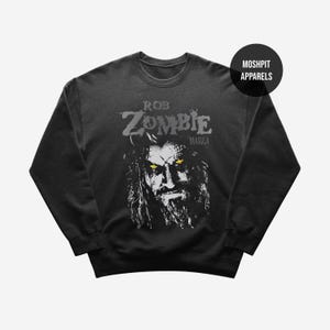 May include: Dark gray sweatshirt featuring a black and white graphic of Rob Zombie with yellow eyes and the text "Rob Zombie Dragula". The sweatshirt has a crew neck and long sleeves. The brand logo "Moshpit Apparels" is on the upper right.