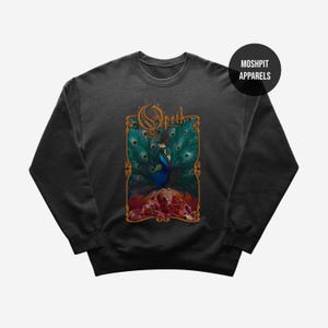 Puede incluir: Sudadera gris oscuro con un diseño de pavo real y la palabra "Opeth" en una fuente adornada. El pavo real tiene plumas azules y verdes, y el diseño incluye un borde floral. La marca "Moshpit Apparels" está en un círculo negro.