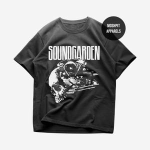 Puede incluir: Camiseta gris oscuro con un gráfico blanco que presenta el nombre de la banda "SOUNDGARDEN" sobre un diseño de calavera y motor. La camiseta tiene cuello redondo y mangas cortas. El logotipo de la marca "MOSHPIT APPARELS" está en la parte superior derecha.