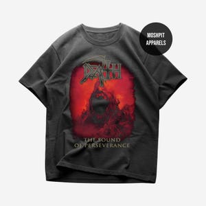 Puede incluir: Camiseta gris oscuro con la palabra "DEATH" en rojo y verde, con un fondo rojo y una figura. El texto "THE SOUND OF PERSEVERANCE" está debajo del gráfico. La camiseta tiene cuello redondo y mangas cortas. La marca "MOSHPIT APPARELS" está arriba a la derecha.