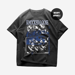 Puede incluir: Camiseta gris oscuro con el texto "KNOCKED LOOSE" en blanco, sobre un diseño gráfico azul y blanco. La camiseta también presenta el texto "A DIFFERENT SHADE OF BLUE" en azul. Un círculo negro con "MOSHPIT APPARELS" está en la parte superior derecha.