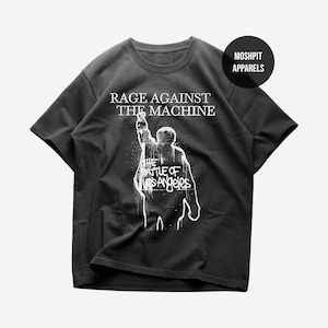 Puede incluir: Camiseta gris oscuro con el texto "RAGE AGAINST THE MACHINE" sobre un gráfico blanco de una persona con el brazo levantado. Debajo, el texto "THE BATTLE OF LOS ANGELES". La camiseta tiene cuello redondo y mangas cortas.